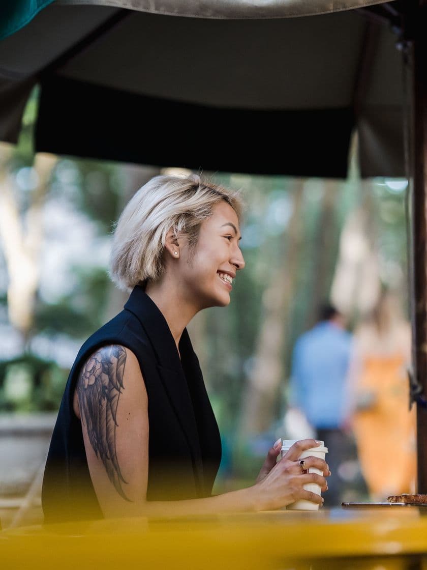 A woman with a tattooed arm smiling over coffee at an outdoor table