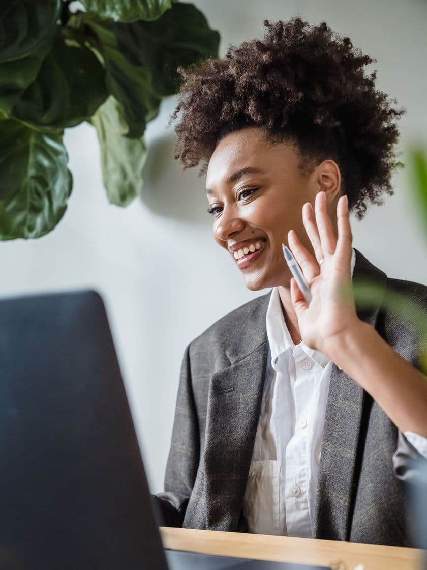 A woman waving hello to a video call at her desk
