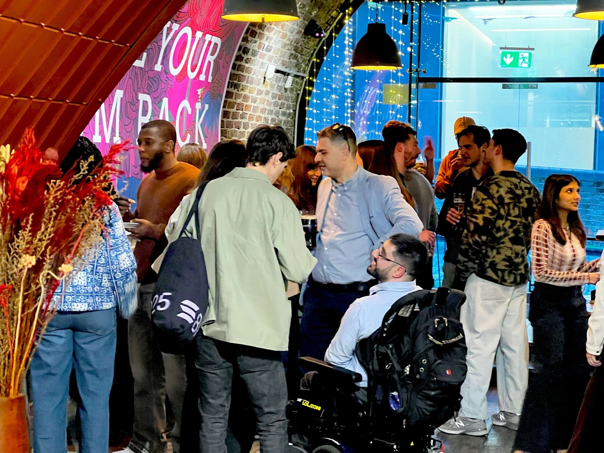 Guests at the Flynt meetup mingle in tight clusters near the venue entrance, including a wheelchair user in conversation with two others. The "Phone Your Mum Back" mural and a fairy-lit doorway glow in the background.