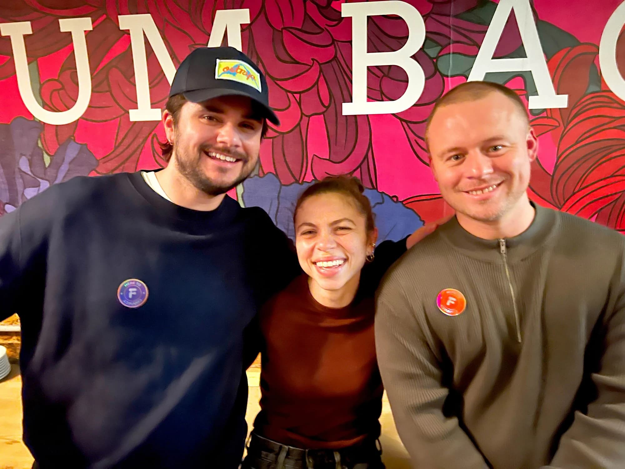 Three Flynt members pose for a group photo in front of the venue's red floral mural. Two wear round holographic "Here to Connect" Flynt badges, all three smiling warmly.