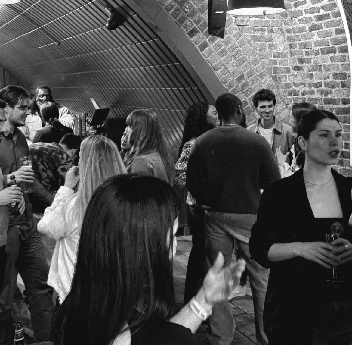 Black and white photo of a Flynt meetup: attendees mingling and chatting in small groups under the arched brick ceiling of a London railway arch venue, drinks in hand, warm pendant lighting overhead.