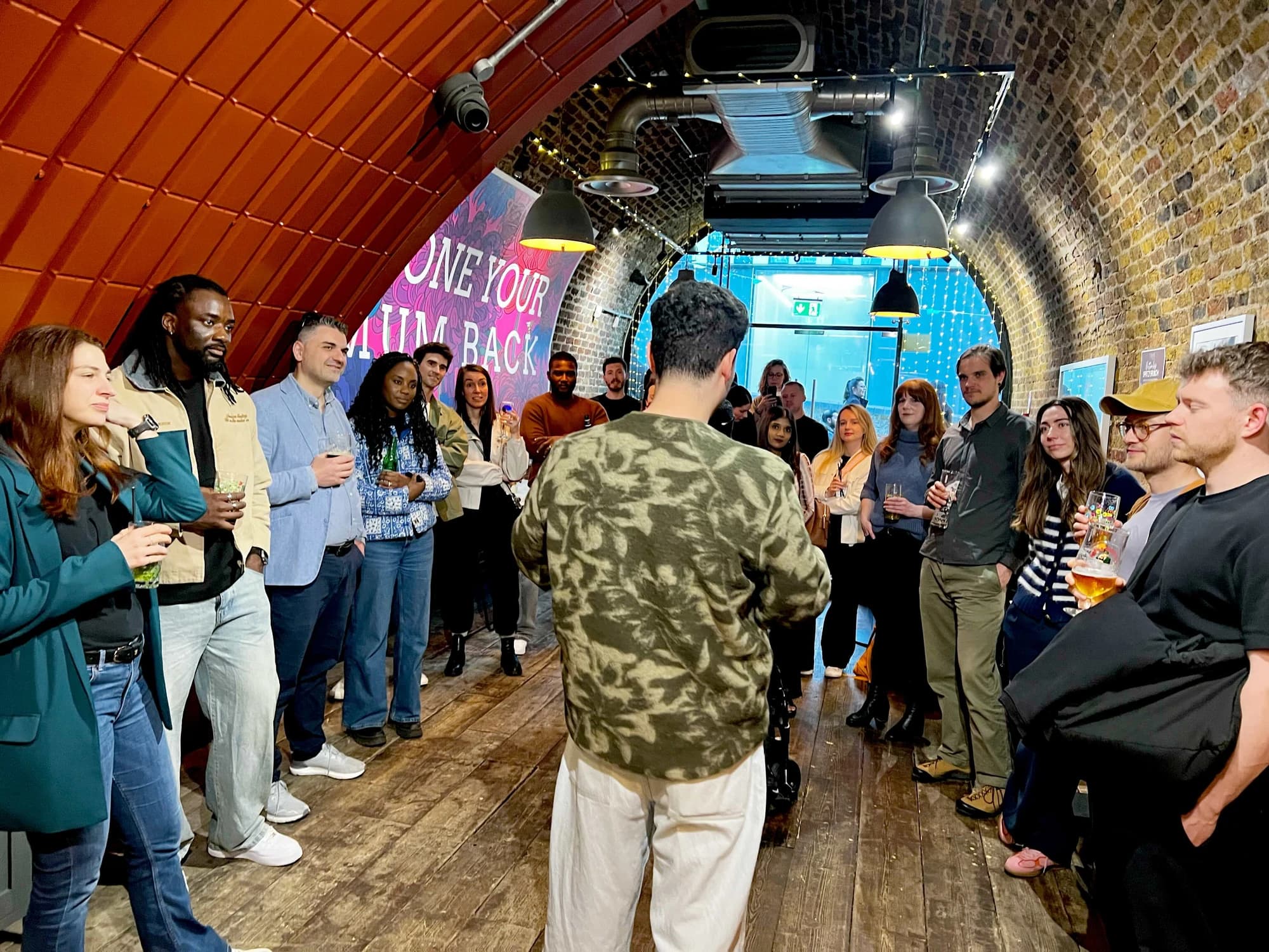 A Flynt host in a camo-print sweatshirt addresses a semicircle of guests inside a brick-arched venue with fairy lights and a bold floral mural reading "Phone Your Mum Back" in the background. Attendees hold drinks and listen.