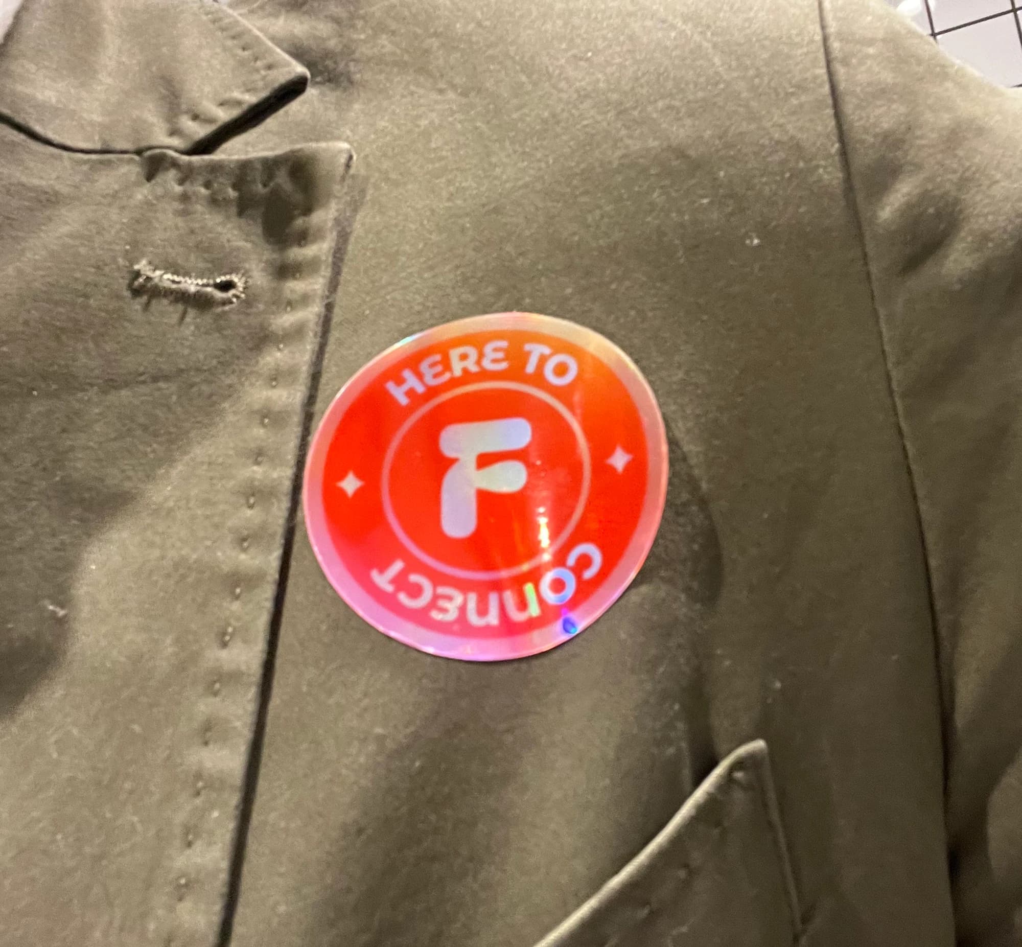 Close-up of a round holographic Flynt badge pinned to an olive jacket lapel, reading "Here to Connect" around the Flynt "F" monogram.